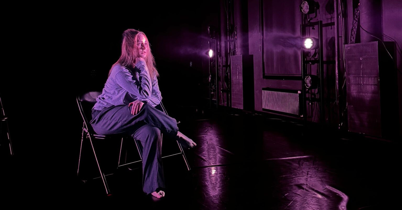 Pitkätukkainen, löysään vaatetukseen pukeutunut henkilö istuu paljain jaloin tuolilla pimeällä näyttämöllä, joka on valaistu violetilla valaistuksella, kädet ristissä ja mietteliäs ilme.