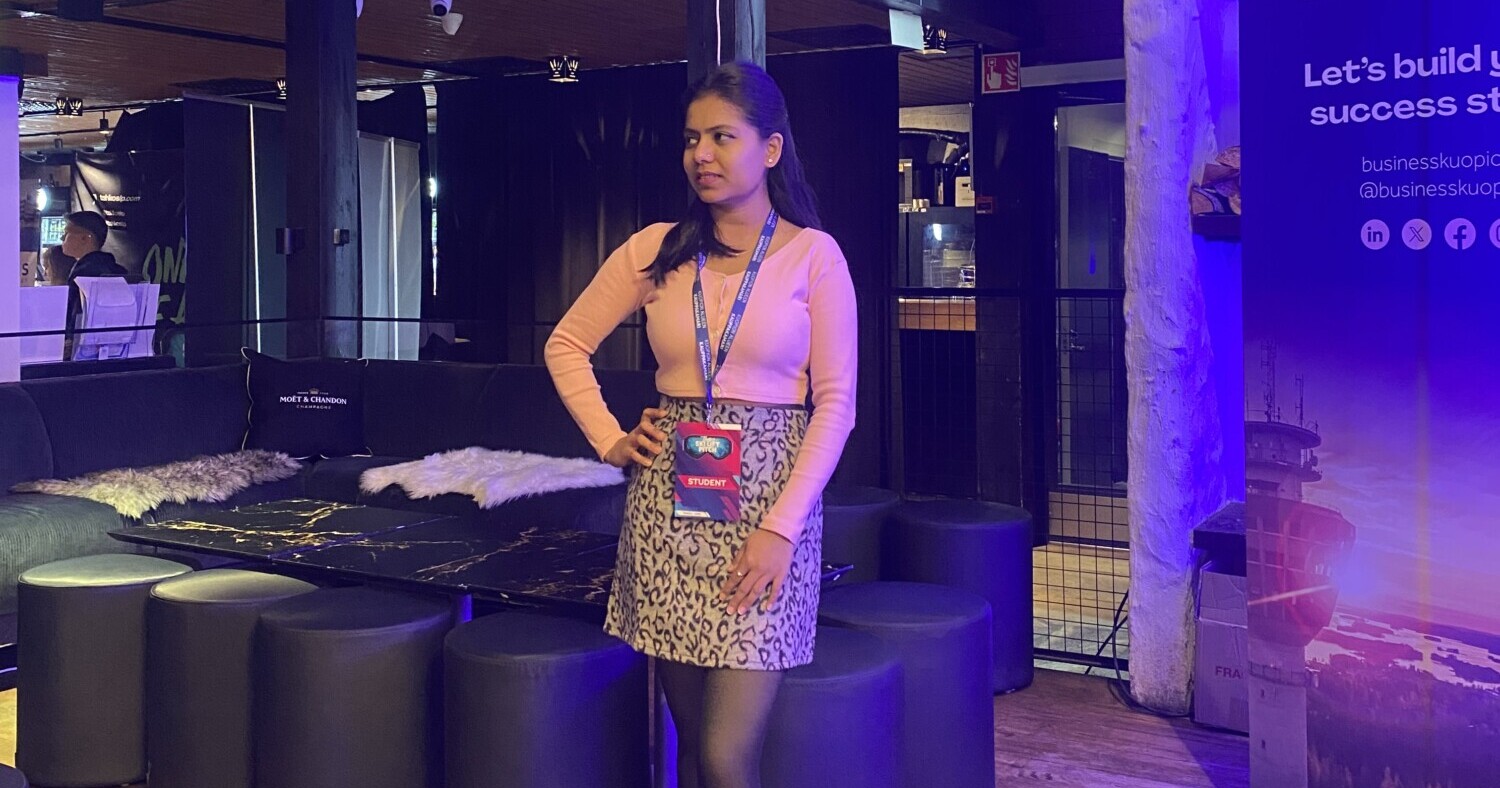 Nainen seisoo sisätiloissa mustan sohvan vieressä, yllään vaaleanpunainen toppi, kuviollinen hame ja tapahtumamerkki. Taustalla on oleskeluhuonekaluja ja violetti banneri, jossa on yritystukitietoja.