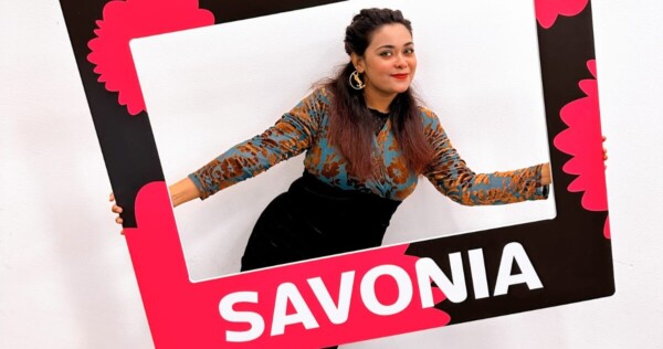 Nainen poseeraa leikkisästi suuren, mustan, vaaleanpunaisen ja valkoisen suorakulmaisen kehyksen sisällä, johon on kirjoitettu sana SAVONIA, valkoista taustaa vasten.