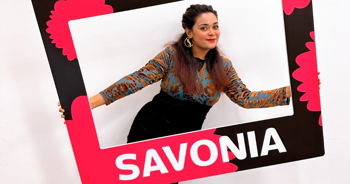 Nainen poseeraa leikkisästi suuren, mustan, vaaleanpunaisen ja valkoisen suorakulmaisen kehyksen sisällä, johon on kirjoitettu sana SAVONIA, valkoista taustaa vasten.