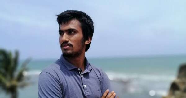 Lyhyet tummat hiukset ja leikattu parta omaava mies, jolla on harmaa poolopaita, seisoo kädet ristissä ja katsoo vasemmalle. Taustalla näkyy sumea rantakuva, jossa on meri ja palmu.