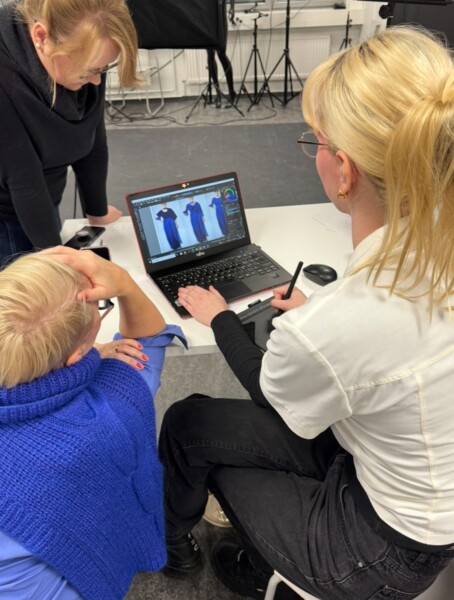 Kolme ihmistä kokoontuu kannettavan tietokoneen ympärille muokkaamaan sinipukuisen henkilön digitaalisia kuvia kynää ja grafiikkataulukkoa käyttäen studiossa, jonka taustalla näkyy valokuvauslaitteita.