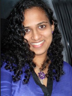 Tummahiuksinen kihara nainen, jolla on violetti toppi ja siihen sopiva kaulakoru, hymyilee kameralle istuessaan sisätiloissa.