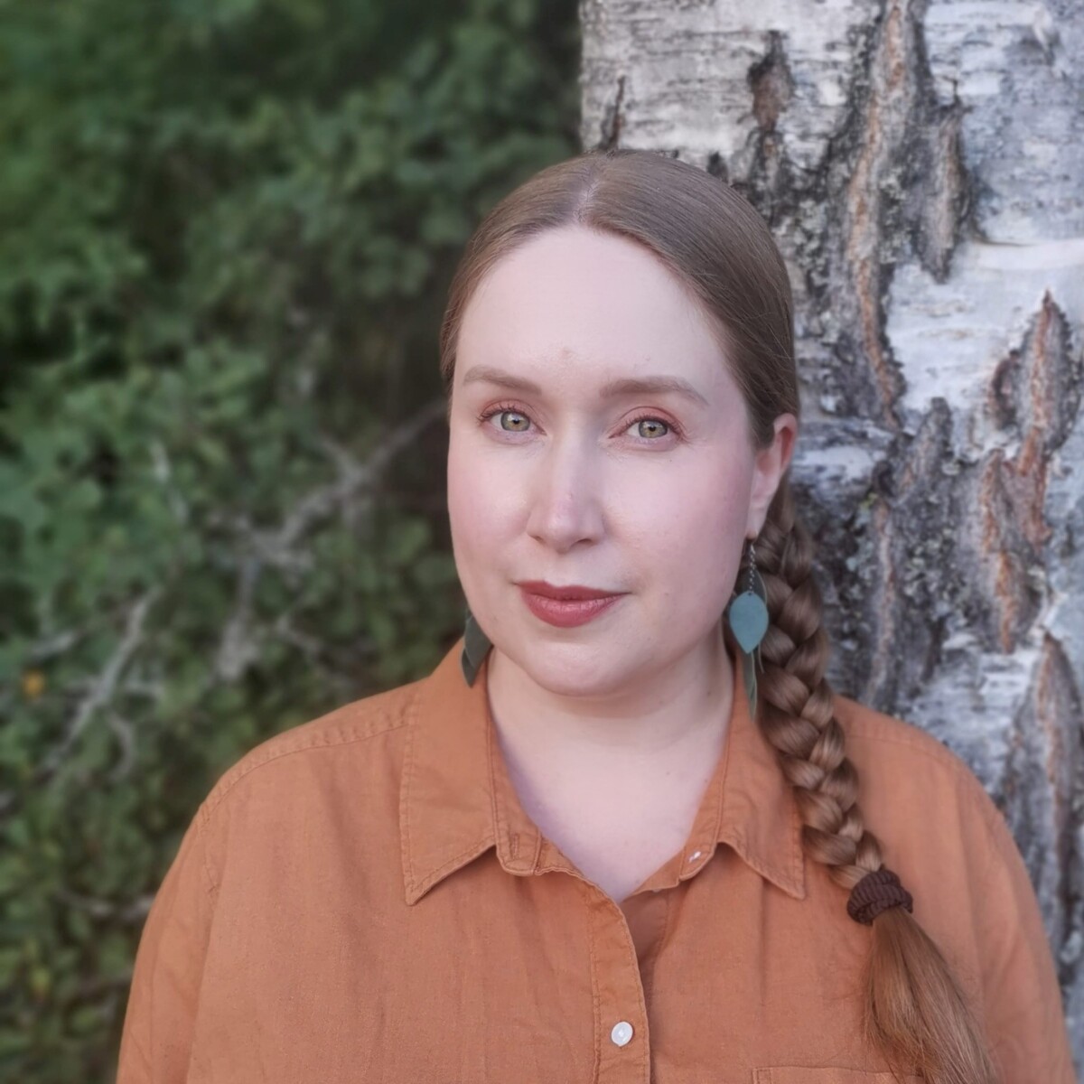 Vaaleaihoinen nainen, jolla on pitkät, vaaleanruskeat hiukset letissä, seisoo ulkona koivun luona. Hänellä on yllään ruosteenvärinen paita ja vihreät korvakorut, ja taustalla on hämärtynyt vehreys.