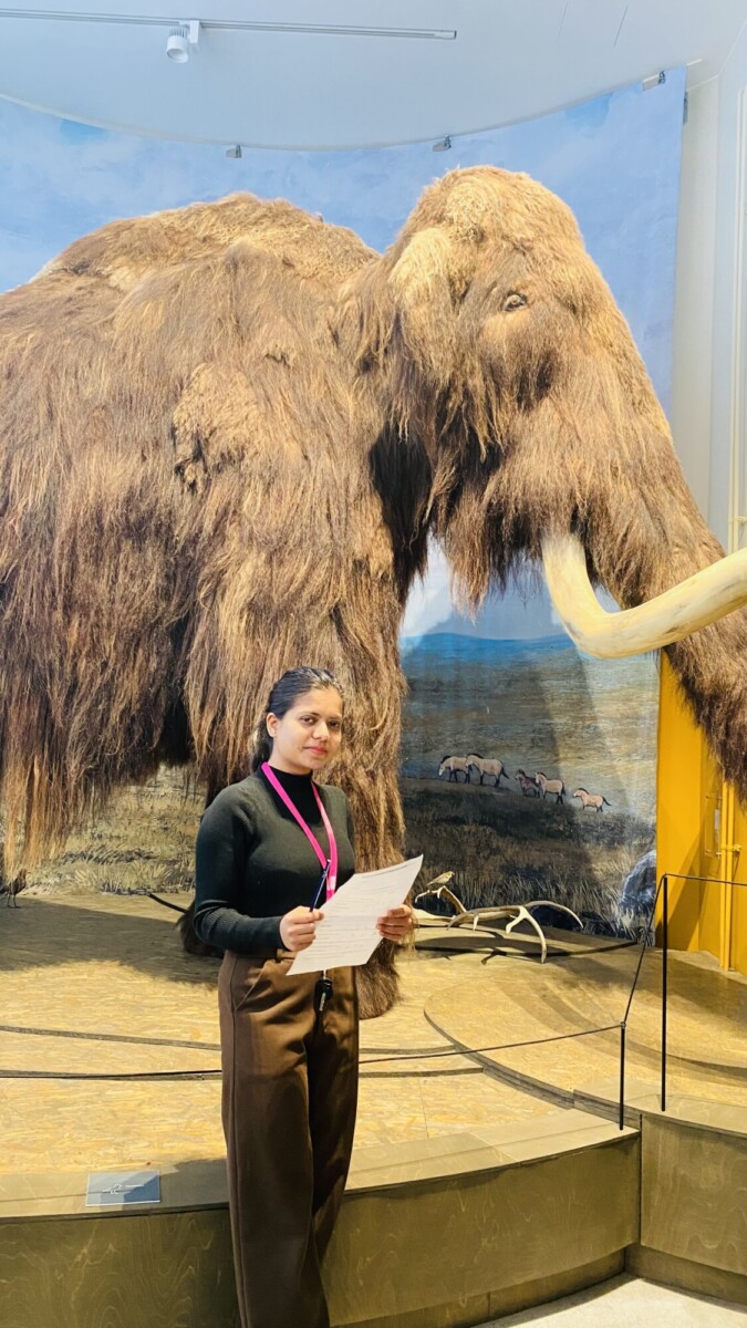 Nainen, jolla on kädessään papereita, seisoo museon suuren villamammutti-näyttelyn edessä. Hänellä on ruskeat housut, musta toppi ja vaaleanpunainen kaulanauha. Taustalla on seinämaalausta ruohoisesta tasangosta, jossa on eläimiä.