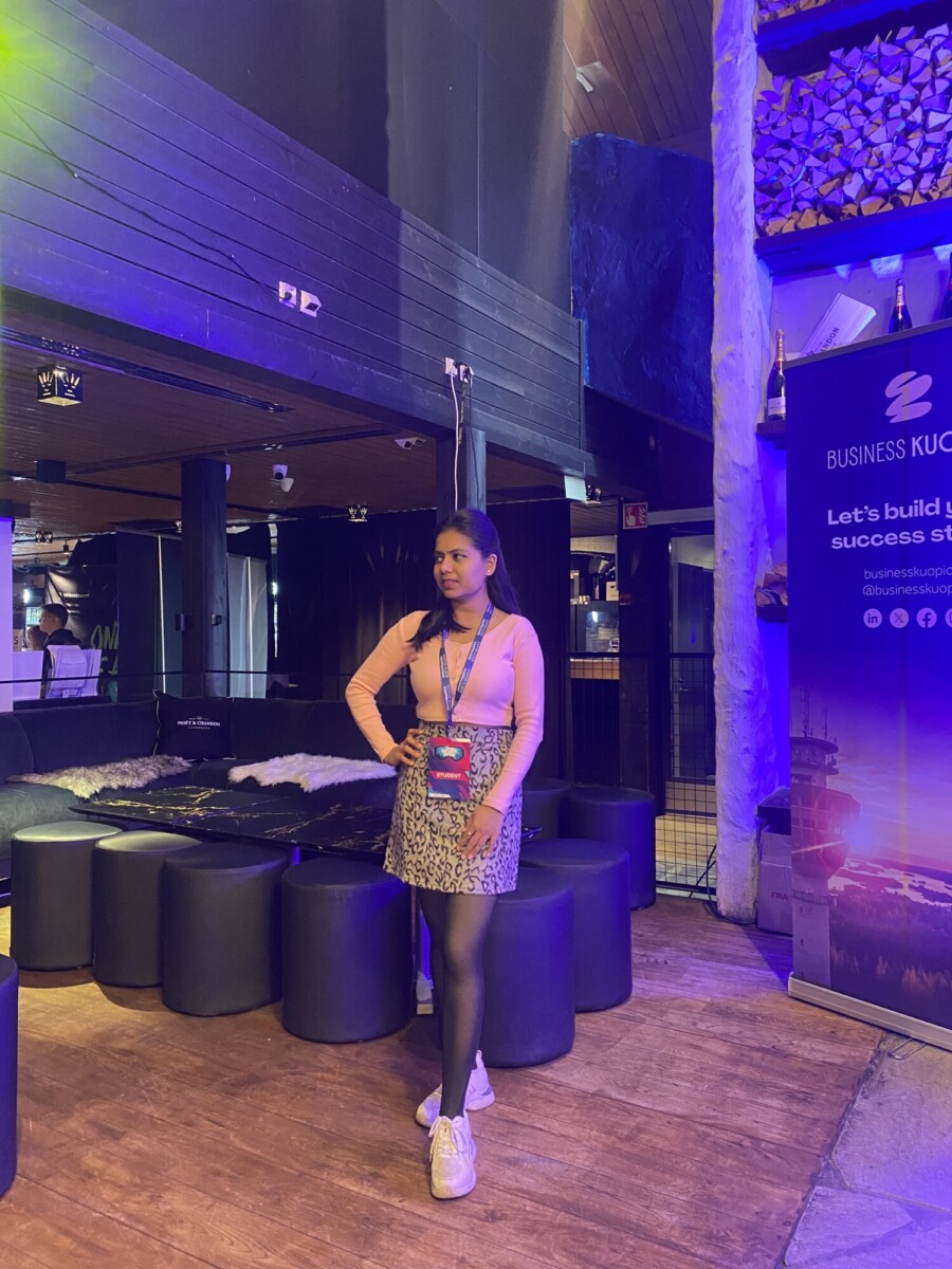 Nainen poseeraa sisätiloissa tapahtumatilassa, jossa on modernit mustat istuimet, ja hänellä on päällään nauhaköysi, vaaleanpunainen toppi ja leopardikuvioinen hame. Oikealla on Business Kuopio -banneri. Valaistus on värikäs ja tunnelmallinen.