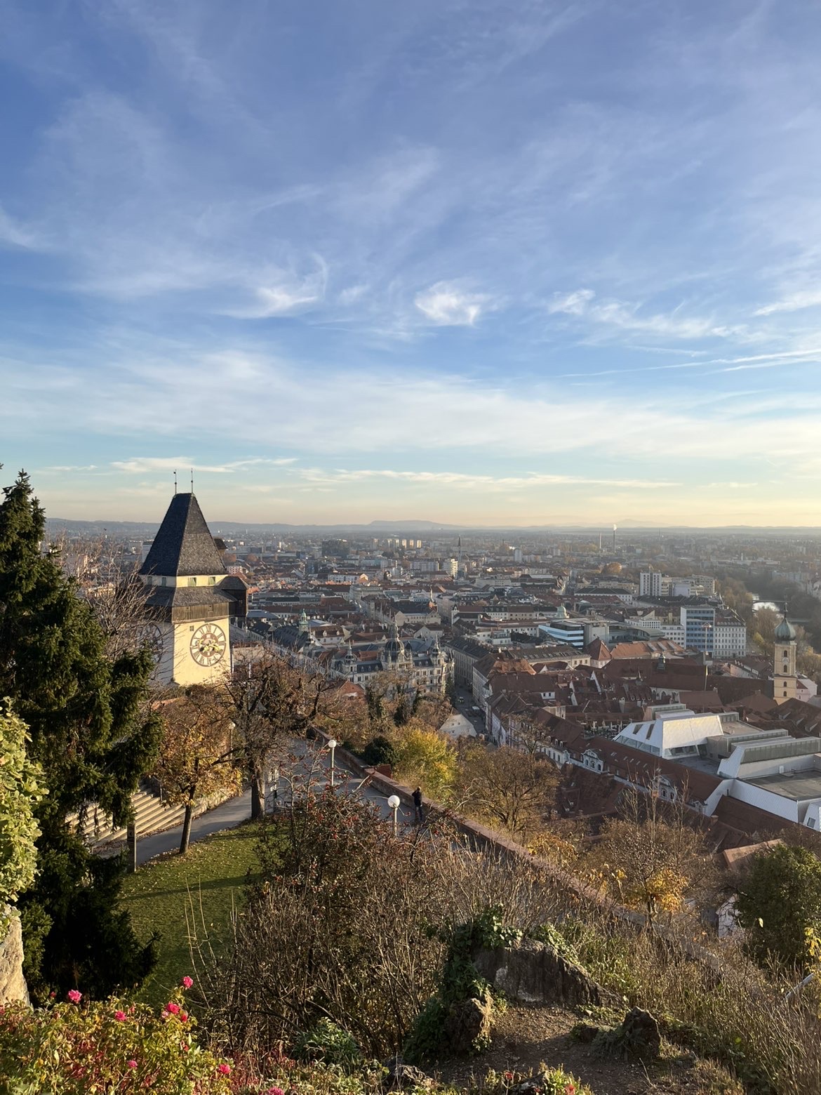 Näkymä Itävallan Grazista, jossa on ikoninen Uhrturm (kellotorni) kukkulalla vehreyden ympäröimänä, ja kaupungin katot ja rakennukset levittäytyvät sinisen taivaan ja pilvien peitossa.