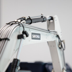 Lähikuva valkoisesta mekaanisesta käsivarresta tai rakennuslaitteen osasta, jossa on näkyvissä kaapeleita ja SAVONIA-etiketti, tavallisella, sumealla taustalla.