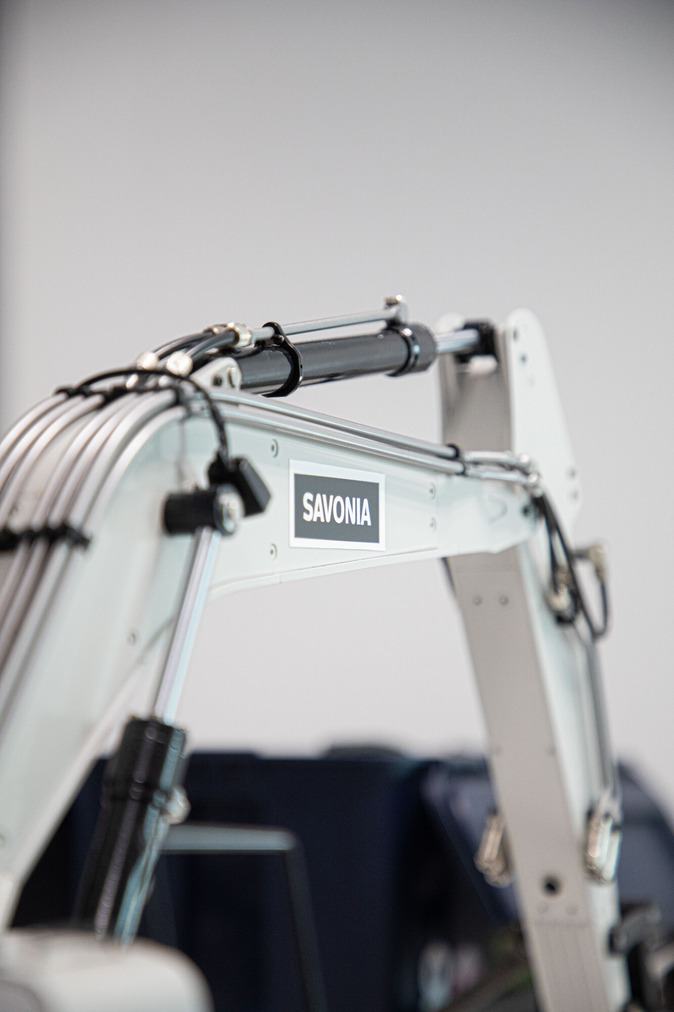 Lähikuva valkoisesta mekaanisesta käsivarresta tai rakennuslaitteen osasta, jossa on näkyvissä kaapeleita ja SAVONIA-etiketti, tavallisella, sumealla taustalla.