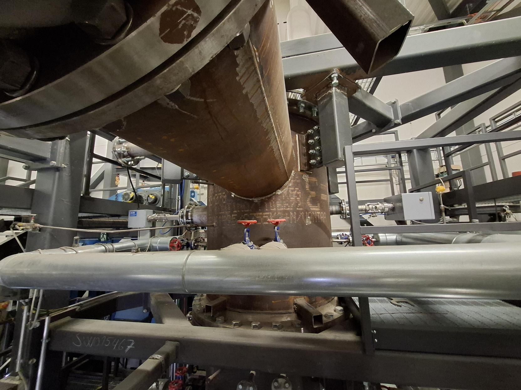 Suuria teollisuusputkia ja metallirakenteita tehdasympäristössä, jossa on paksu ruosteenvärinen pystysuora putki yhdistettynä vaakasuoriin hopeisiin putkiin ja punaisiin venttiileihin. Taustalla näkyy lisää laitteita ja rakennepalkkeja.