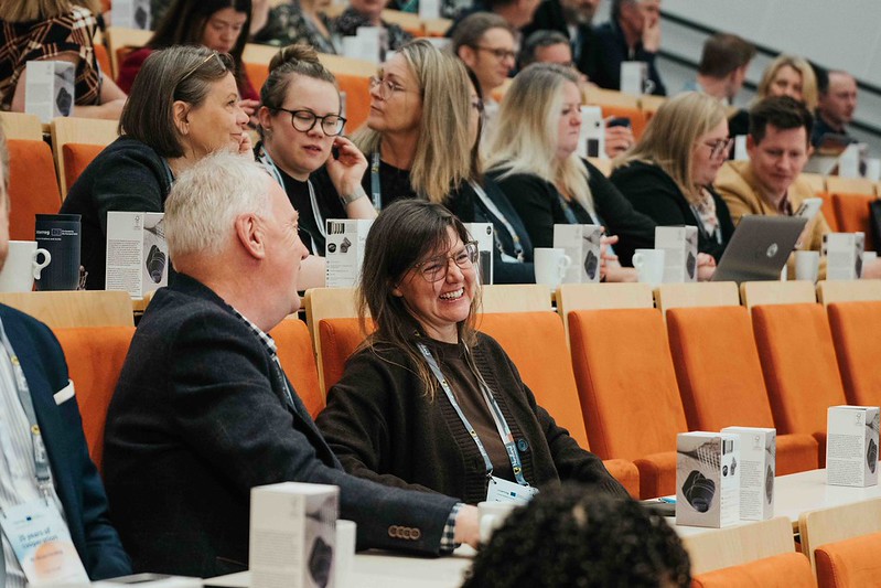 Ihmiset istuvat oransseissa tuolirivissä konferenssissa tai seminaarissa. Etualalla kaksi ihmistä hymyilee ja puhuu, kun muut kuuntelevat, käyttävät kannettavia tietokoneita tai keskustelevat. Kahvikupit ja laatikot ovat heidän edessään olevilla pöydillä.