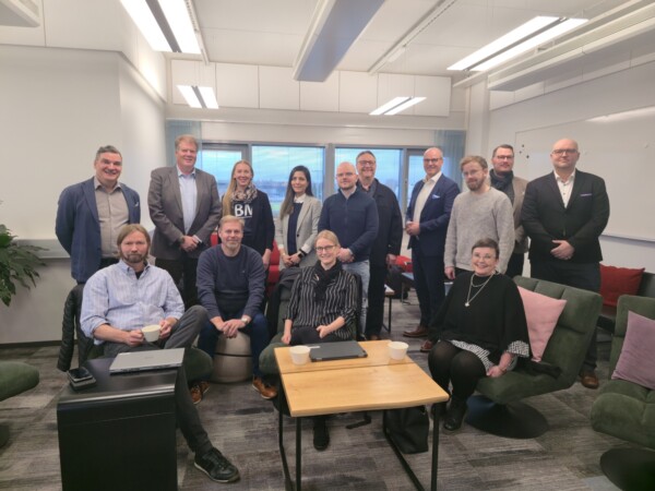 Viidentoista ihmisen ryhmä, sekä miehiä että naisia, poseeraa ja hymyilee toimiston kokoushuoneessa. Jotkut istuvat puupöydän ympärillä kannettavien tietokoneiden ja kahvikuppien kanssa, kun taas toiset seisovat heidän takanaan. Tunnelma on rento ja ystävällinen.