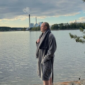 Harmaaseen kylpytakkiin pukeutunut mies seisoo kivikkoisella rannalla ja katselee pilvisen taivaan alla rauhalliselle järvelle, jonka kaukaisuudessa on tehdas ja savupiippu. Oikealla näkyy männyn oksia.