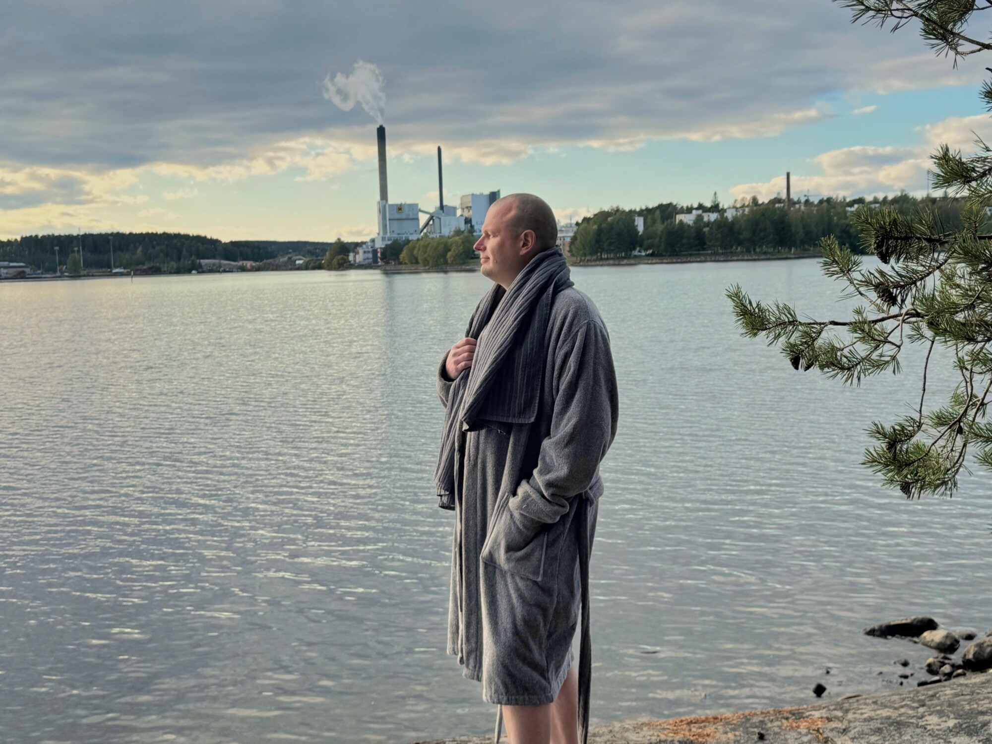 Harmaaseen kylpytakkiin pukeutunut mies seisoo kivikkoisella rannalla ja katselee pilvisen taivaan alla rauhalliselle järvelle, jonka kaukaisuudessa on tehdas ja savupiippu. Oikealla näkyy männyn oksia.