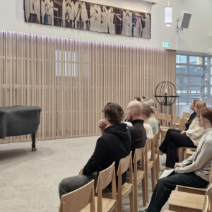 Mies seisoo katetun flyygelin vieressä ja puhuu istuvalle yleisölle modernissa huoneessa, jossa on puinen sisustus ja suuret ikkunat. Yleisö kuuntelee tarkkaavaisesti.