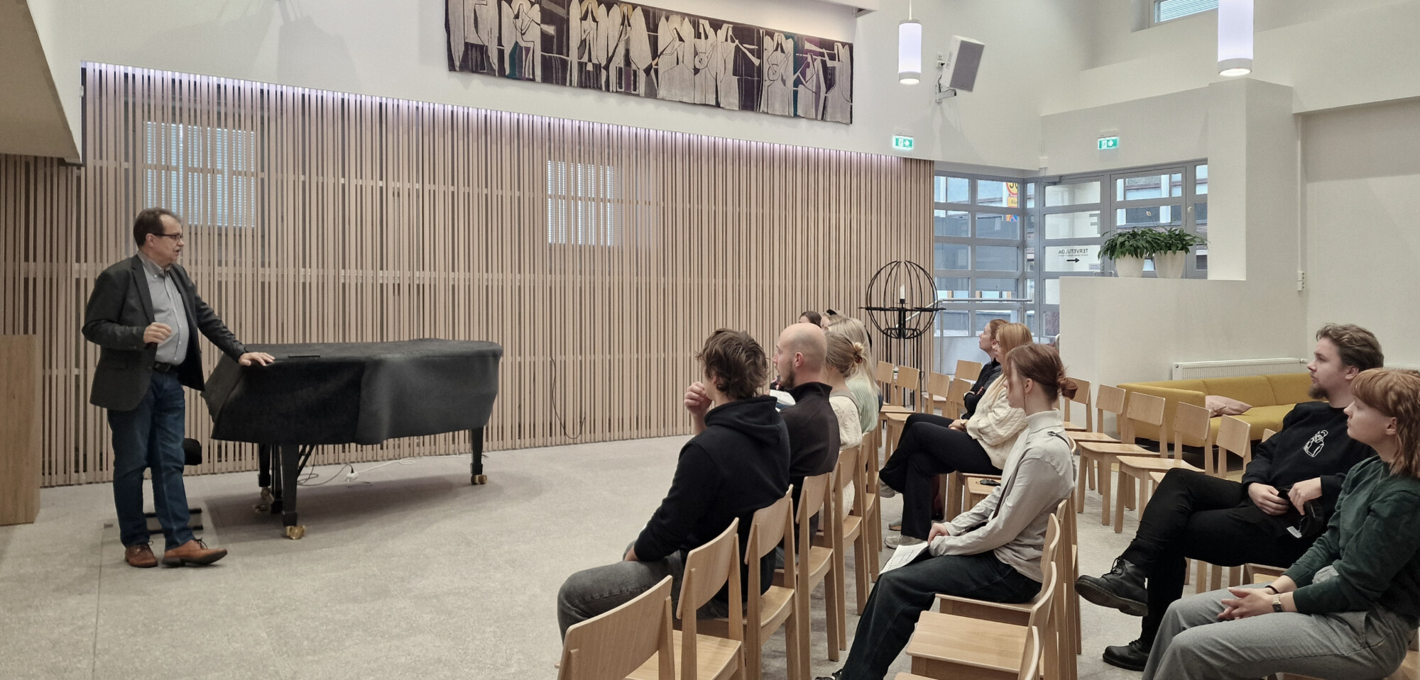 Mies seisoo katetun flyygelin vieressä ja puhuu istuvalle yleisölle modernissa huoneessa, jossa on puinen sisustus ja suuret ikkunat. Yleisö kuuntelee tarkkaavaisesti.