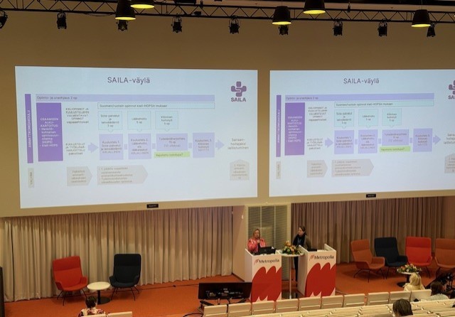 Kaksi esitelmöitsijää seisoo korokkeella modernissa luentosalissa ja puhuu suurten valkokankaiden edessä, joilla näkyy suomenkielinen SAILA-väylä -niminen vuokaavio. Salissa on oranssit tuolit ja vaaleat verhot.