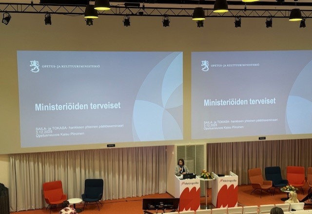 Puhuja seisoo luentosalissa korokkeella ja puhuu yleisölle. Kahdella suurella näytöllä näkyy dia otsikolla Ministeriöiden terveiset, jossa on suomenkielisiä logoja ja tekstiä, ja lavalla on useita tyhjiä tuoleja.