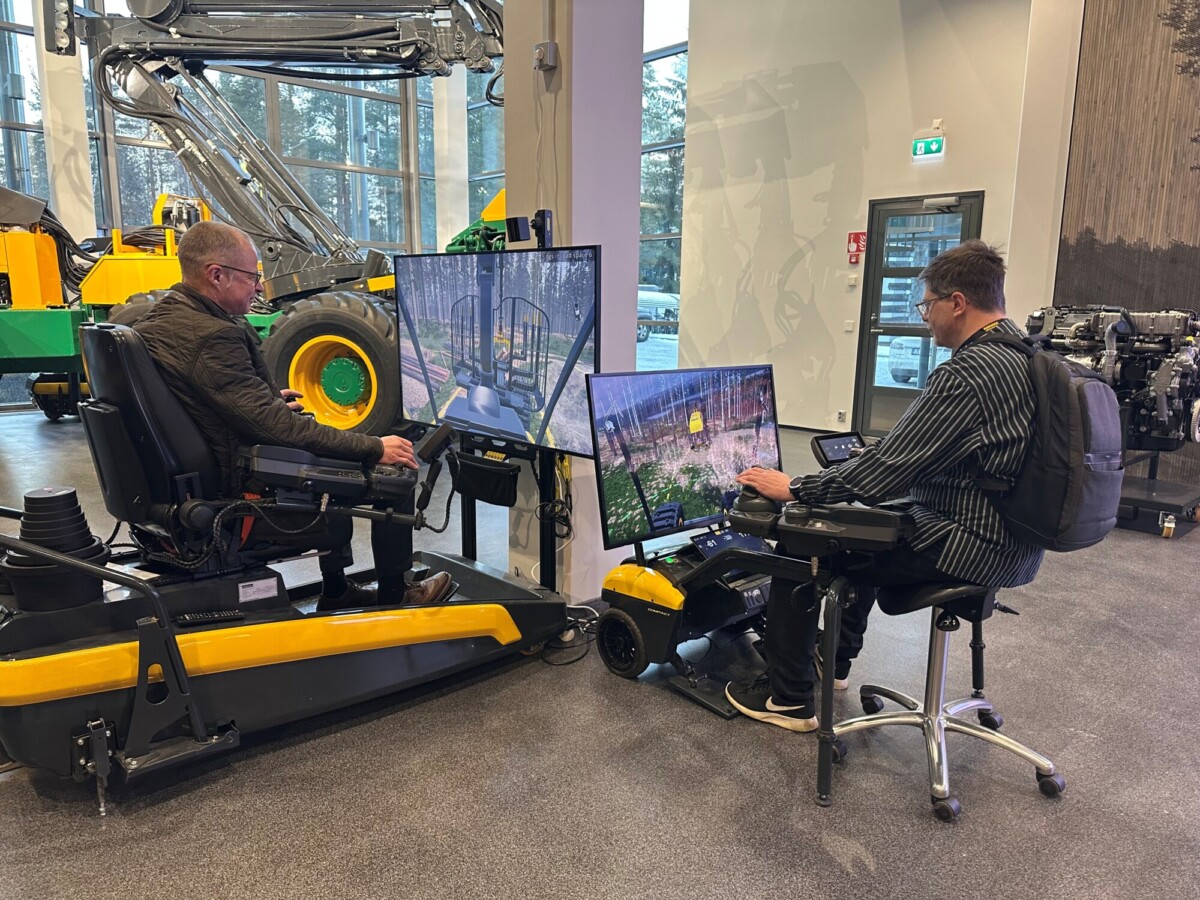 Kaksi miestä istuu suurilla ohjausasemilla, joissa on useita näyttöjä, ja käyttää metsäkonesimulaattoreita modernissa näyttelytilassa, jonka taustalla on esillä koneita ja laitteita.