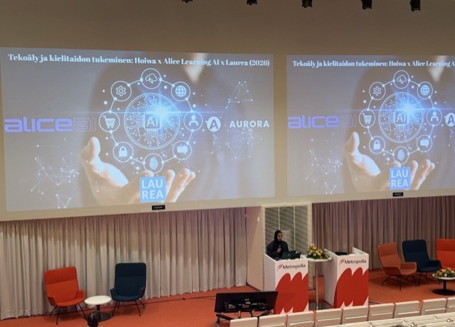 Puhuja seisoo korokkeella modernissa luentosalissa, jossa on punaisia tuoleja. Kahdella suurella näytöllä näkyy esitys tekoälystä ja kielituesta, jossa on logoja ja digitaalisen verkon grafiikkaa.