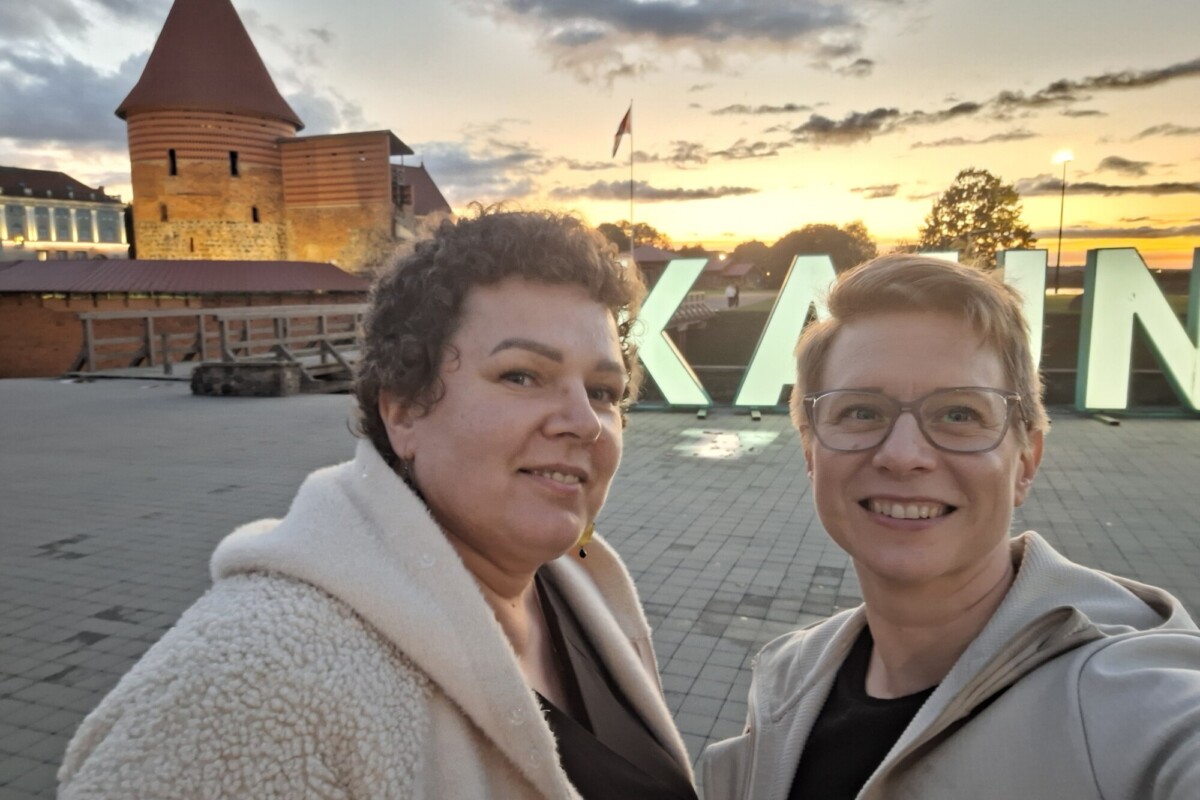 Kaksi hymyilevää ihmistä ottaa selfien ulkona auringonlaskun aikaan, taustalla historiallinen tiilirakennus ja suuret valaistut kirjaimet. Taivas on osittain pilvinen ja illan valo on lämmin.