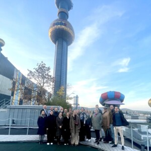 Joukko ihmisiä poseeraa ulkona katolla, ja taustalla näkyy Wienin Spittelaun jätteenpolttolaitoksen värikäs, taiteellinen torni sinisellä taivaalla.