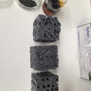 Kolme huokoista, harmaata vaahtomuovipalikkaa, joissa on erilainen sisäinen rakenne, on sijoitettu pystysuoraan valkoiselle pinnalle. Lähellä on lasipurkki, jossa on mustaa materiaalia, pieni pullo, jossa on ruskeaa nestettä, ja nitriilikäsinepakkaus.