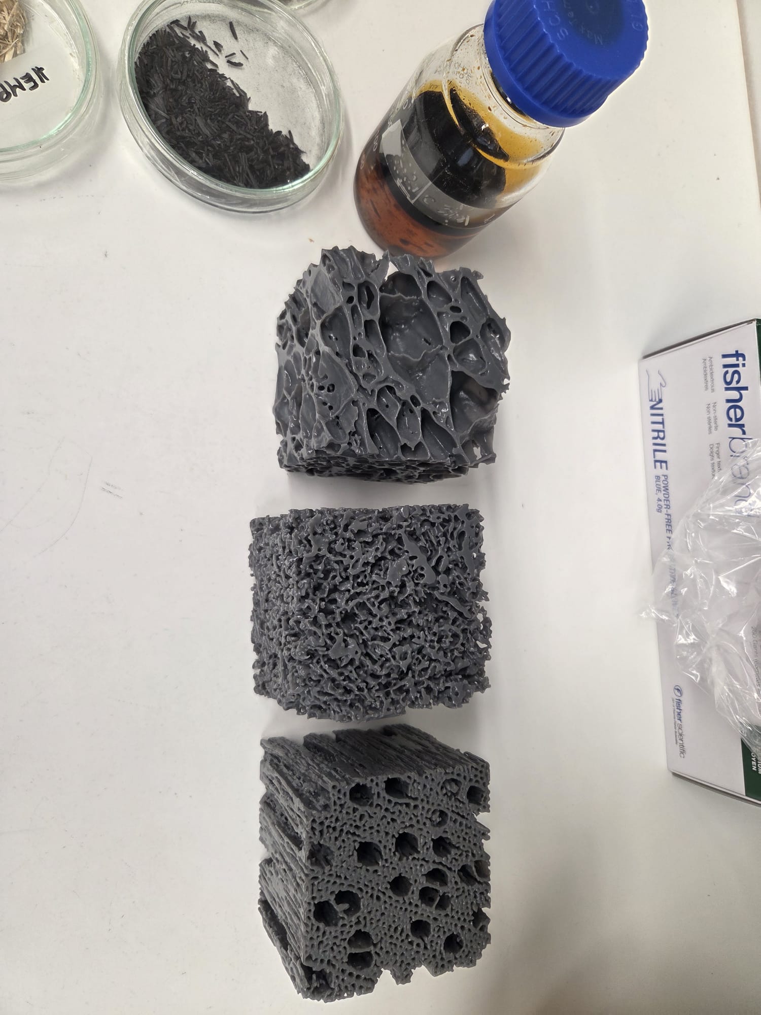 Kolme huokoista, harmaata vaahtomuovipalikkaa, joissa on erilainen sisäinen rakenne, on sijoitettu pystysuoraan valkoiselle pinnalle. Lähellä on lasipurkki, jossa on mustaa materiaalia, pieni pullo, jossa on ruskeaa nestettä, ja nitriilikäsinepakkaus.