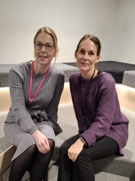 Kaksi naista istuu vierekkäin matolla päällystetyillä portailla ja hymyilee kameralle. Toisella on harmaa mekko ja silmälasit, toisella violetti pusero. Molemmilla on vaaleanruskeat hiukset taakse sidottuina ja heillä on kaulanauhat.