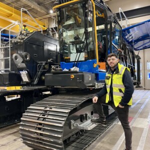 Henkilö, jolla on keltainen turvaliivi ja silmälasit, seisoo suuren, sinimustan telaketjullisen teollisuusajoneuvon vieressä tilavassa varastossa, jossa on korkeat katot ja teollisuuslaitteet.