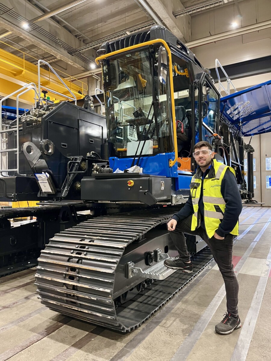 Henkilö, jolla on keltainen turvaliivi ja silmälasit, seisoo suuren, sinimustan telaketjullisen teollisuusajoneuvon vieressä tilavassa varastossa, jossa on korkeat katot ja teollisuuslaitteet.