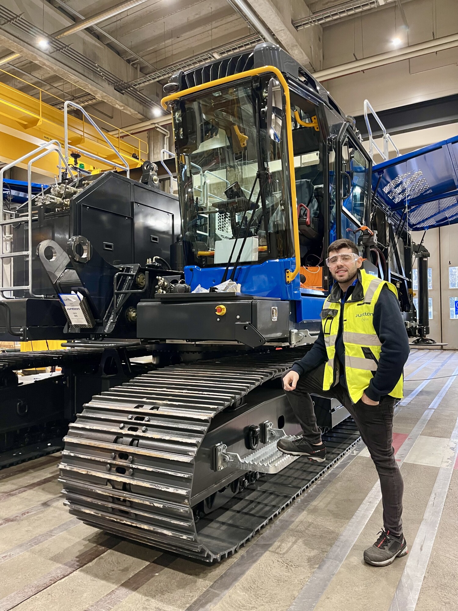 Henkilö, jolla on keltainen turvaliivi ja silmälasit, seisoo suuren, sinimustan telaketjullisen teollisuusajoneuvon vieressä tilavassa varastossa, jossa on korkeat katot ja teollisuuslaitteet.