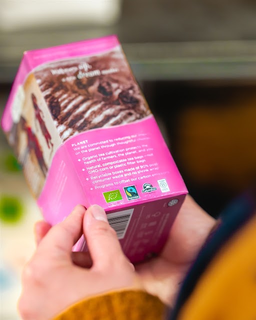 Henkilö pitää kädessään vaaleanpunaista jäätelörasiaa ja keskittyy sen takapuolelle, jossa on ympäristöystävällisyys- ja sertifiointitarroja, ympäristösitoumusteksti ja osa jälkiruoan kuvasta.