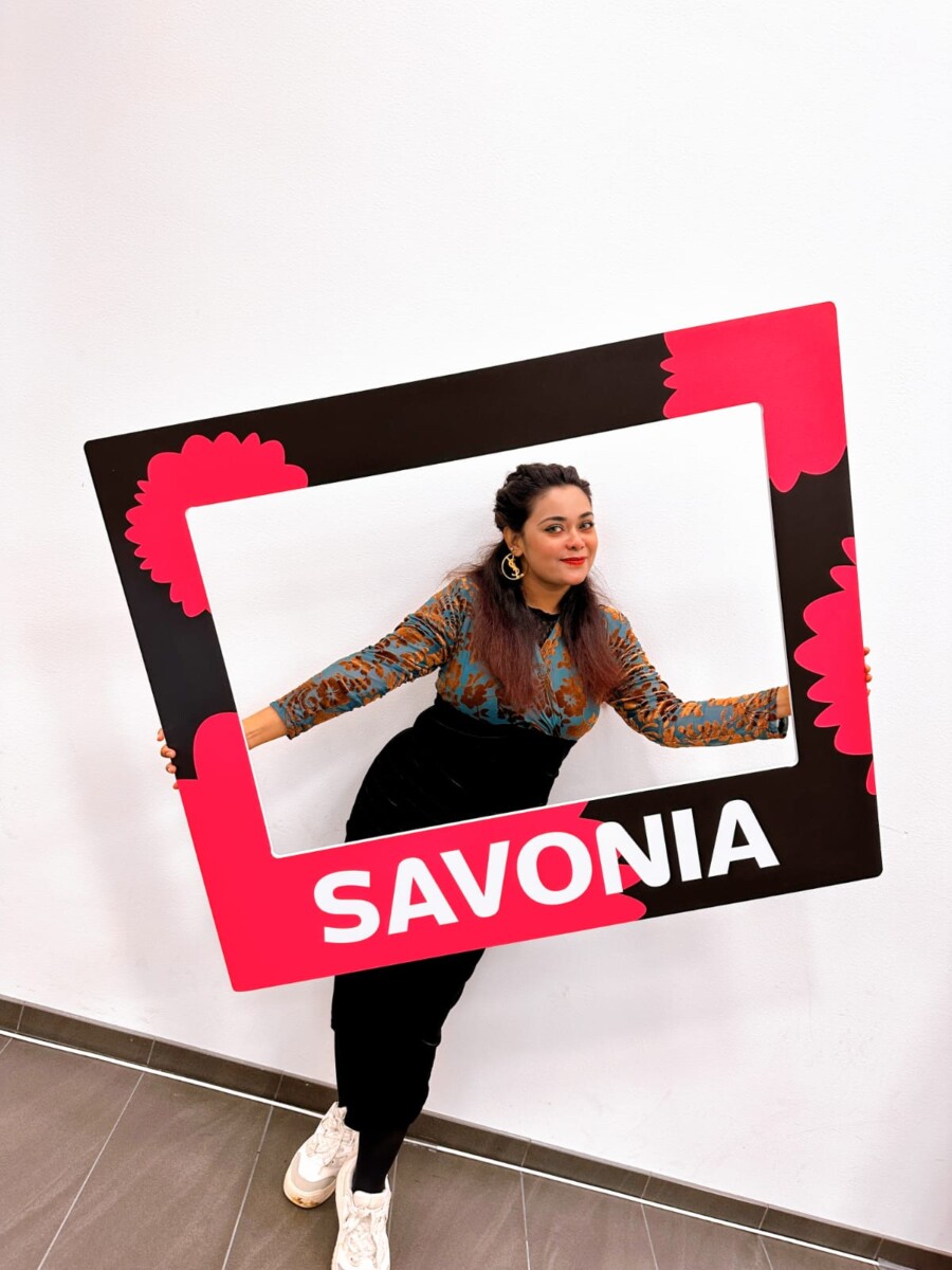 Nainen poseeraa sisätiloissa pitäen kädessään suurta suorakulmaista kehystä, jossa on punaisia kukkia ja alareunassa vaaleanpunaisella pohjalla rohkeilla valkoisilla kirjaimilla kirjoitettu sana SAVONIA. Nainen hymyilee ja seisoo tavallista valkoista seinää vasten.