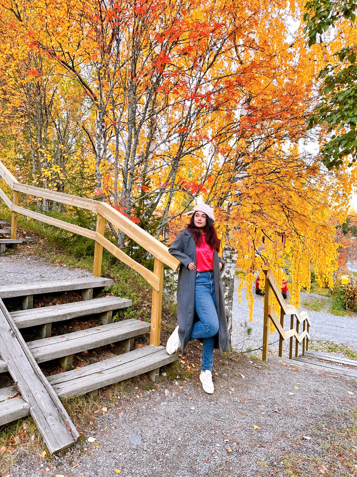 Harmaaseen takkiin, punaiseen toppiin, farkkuihin ja valkoisiin kenkiin pukeutunut nainen poseeraa puuportaiden äärellä elinvoimaisten syyspuiden, joissa on oransseja ja keltaisia lehtiä, vieressä. Kohtauksessa on kirkas, värikäs syksyn tunnelma.