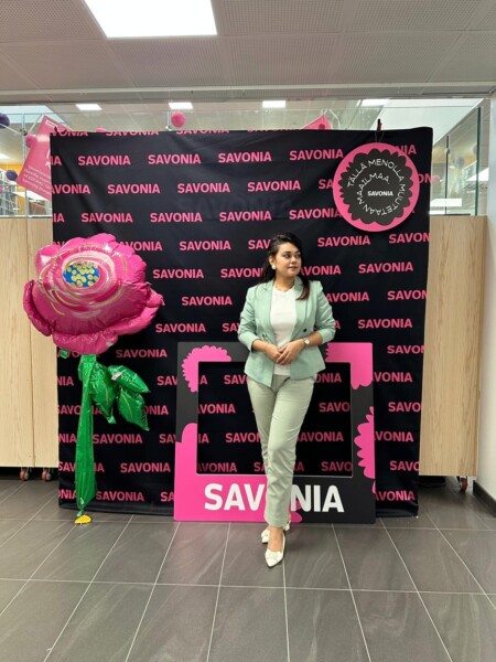 Mintunvihreään pukuun pukeutunut nainen seisoo SAVONIA-kuvataustan edessä suuren vaaleanpunaisen koristeellisen kukan vieressä. Ympäristö näyttää olevan sisätiloissa, joissa on moderni, kaakeloitu lattia.