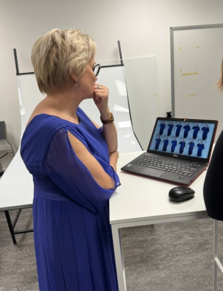 Siniseen mekkoon pukeutunut nainen seisoo korkean työpöydän ääressä ja katsoo mietteliäästi kannettavaa tietokonetta, jossa on kuvia eri asuihin pukeutuneista ihmisistä. Huone on valoisa, ja taustalla on valokuvausstudion laitteita.