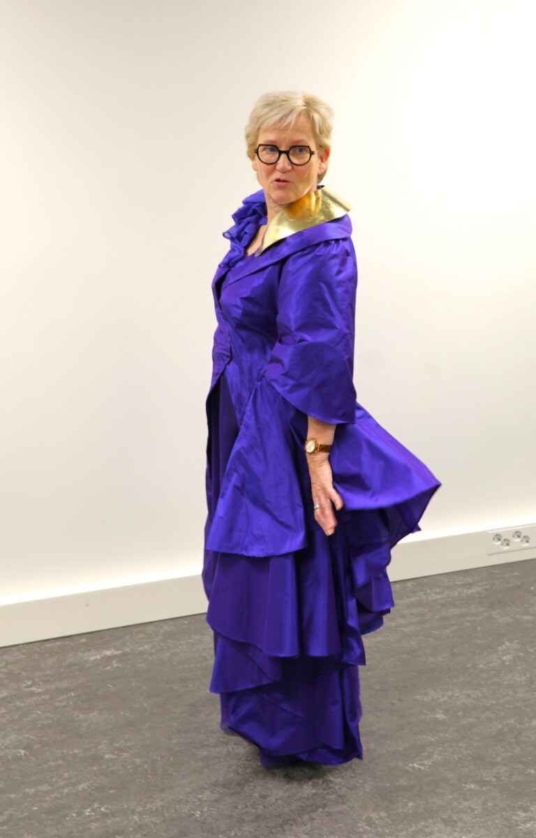 Vanhempi nainen, jolla on silmälasit ja kiiltävä, kerroksellinen violetti puku, poseeraa valkoisen seinän edessä ja katsoo olkansa yli toinen käsi rinnallaan.