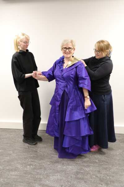 Vanhempi nainen kirkkaan violetissa, kerroksellisessa mekossa seisoo hymyillen, kun kaksi nuorempaa naista auttaa häntä sovittamaan asua. He ovat huoneessa, jossa on tavallinen valkoinen seinä ja harmaa lattia.