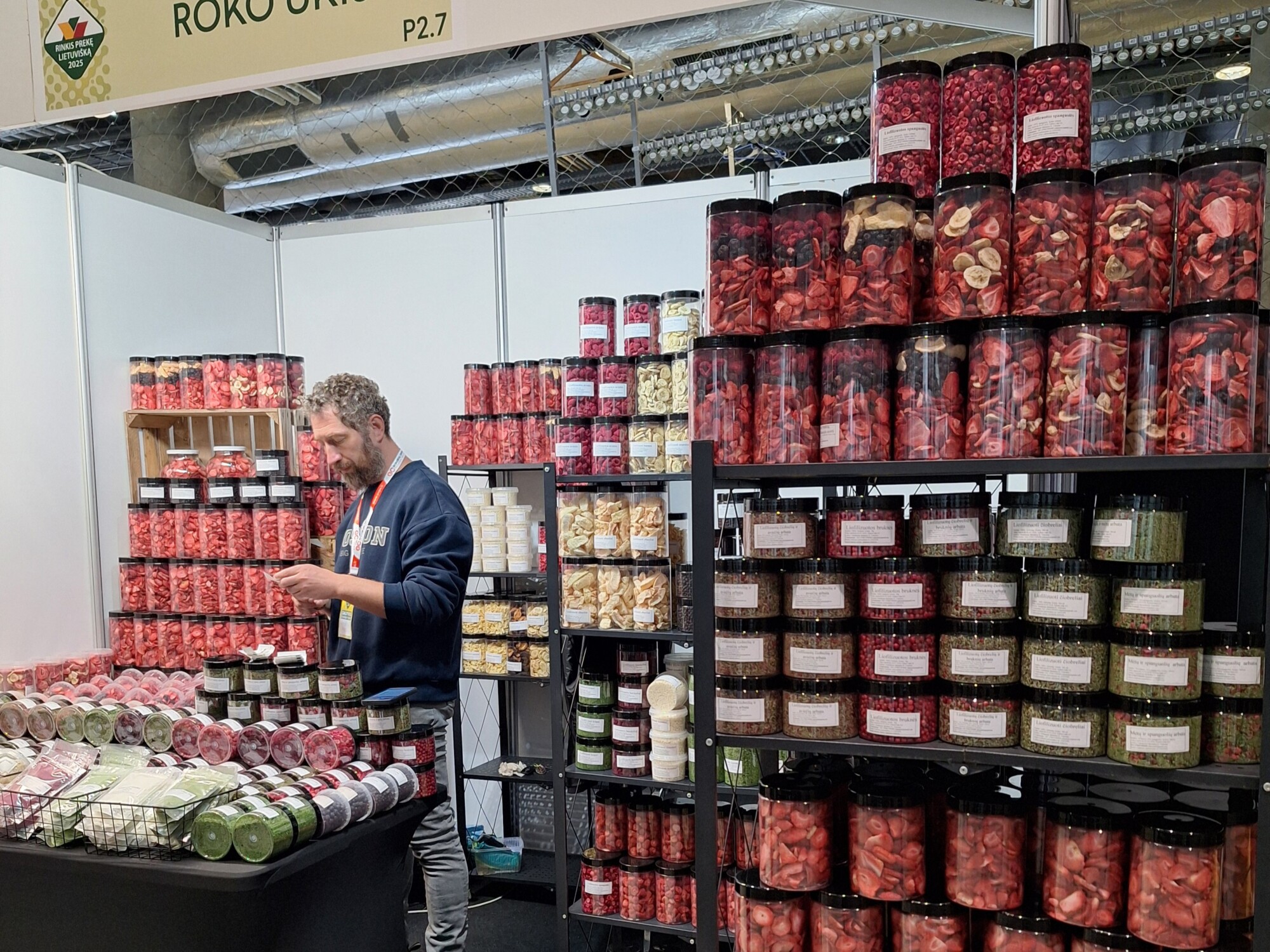 Mies seisoo osastolla, joka on täynnä hyllyjä täynnä värikkäitä purkkeja, jotka sisältävät kuivattuja hedelmiä ja vihanneksia markkinoilla tai messuilla. Purkit on pinottu ja merkitty siististi, ja koju on hyvin valaistu ja järjestetty.