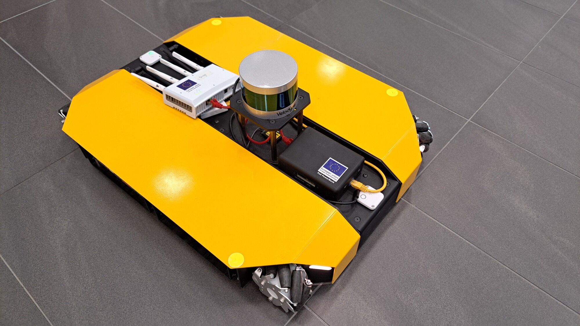 Pieni, suorakaiteen muotoinen keltainen robotti, jonka päällä on antureita ja elektroniikkaa, on sijoitettu harmaalle kaakeloidulle lattialle. Robotin rungon alla on osittain näkyvissä pyörät.