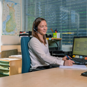 Nainen, jolla on kuulokkeet, istuu työpöydän ääressä ja kirjoittaa muistiinpanovihkoon samalla kun hän katsoo tietokoneen näyttöä. Taustalla näkyy toimistotarvikkeita, asiakirjoja ja seinällä oleva kartta.