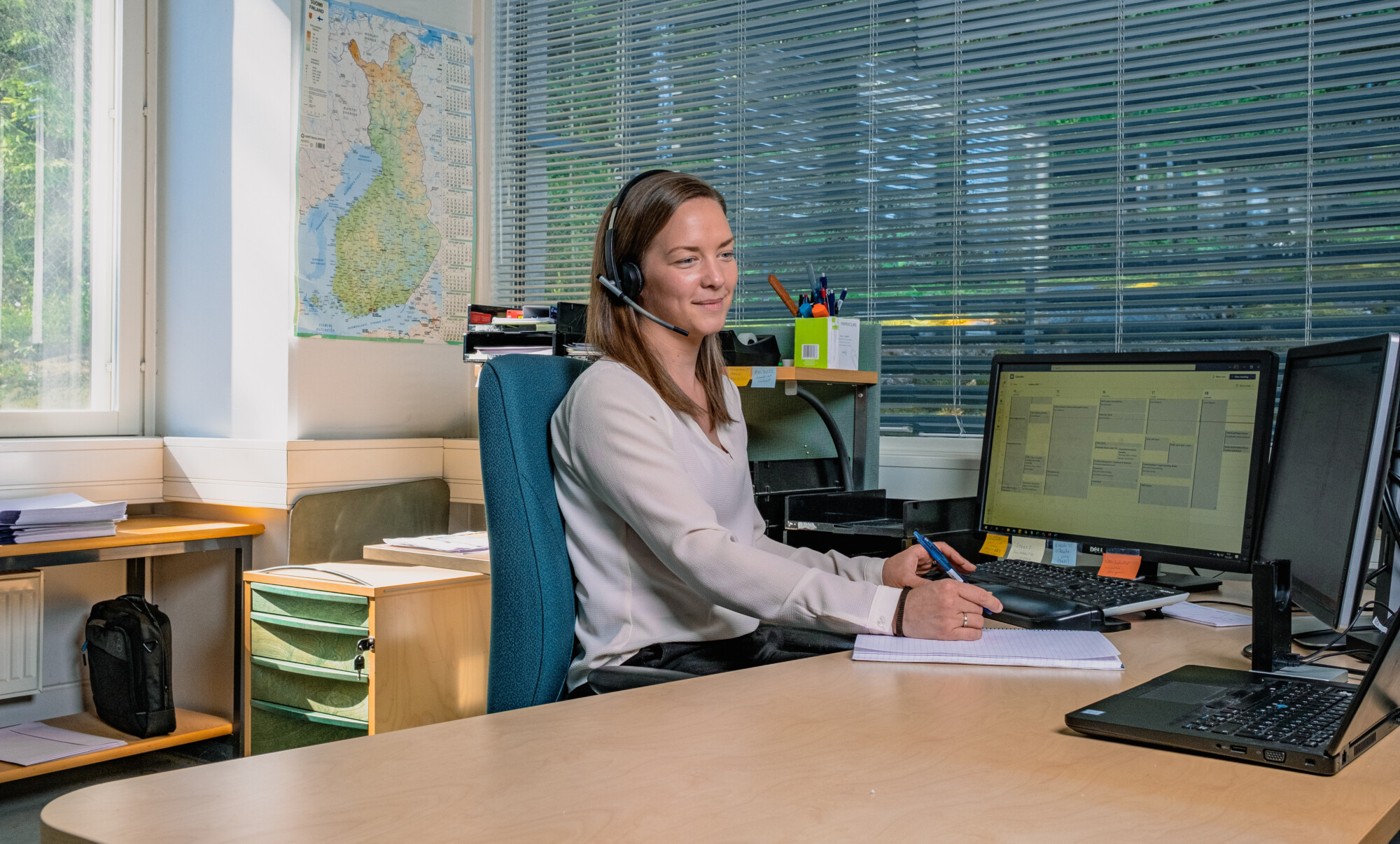 Nainen, jolla on kuulokkeet, istuu työpöydän ääressä ja kirjoittaa muistiinpanovihkoon samalla kun hän katsoo tietokoneen näyttöä. Taustalla näkyy toimistotarvikkeita, asiakirjoja ja seinällä oleva kartta.