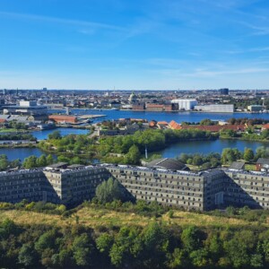 Ilmakuva suuresta, siksak-muotoisesta rakennuksesta, jota ympäröi vehreys ja vesi, taustalla kaupunkikuva ja sininen taivas.