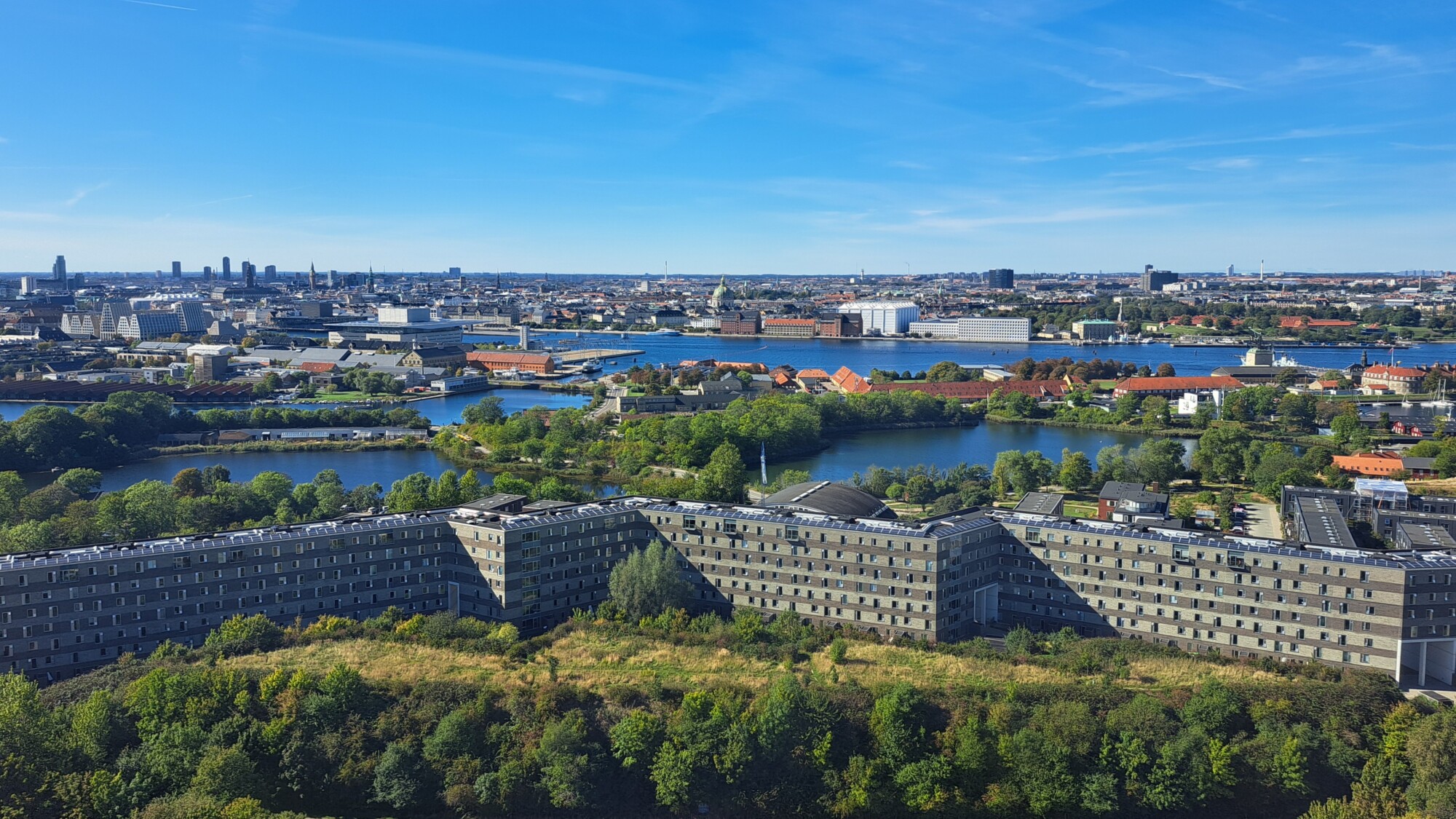 Ilmakuva suuresta, siksak-muotoisesta rakennuksesta, jota ympäröi vehreys ja vesi, taustalla kaupunkikuva ja sininen taivas.