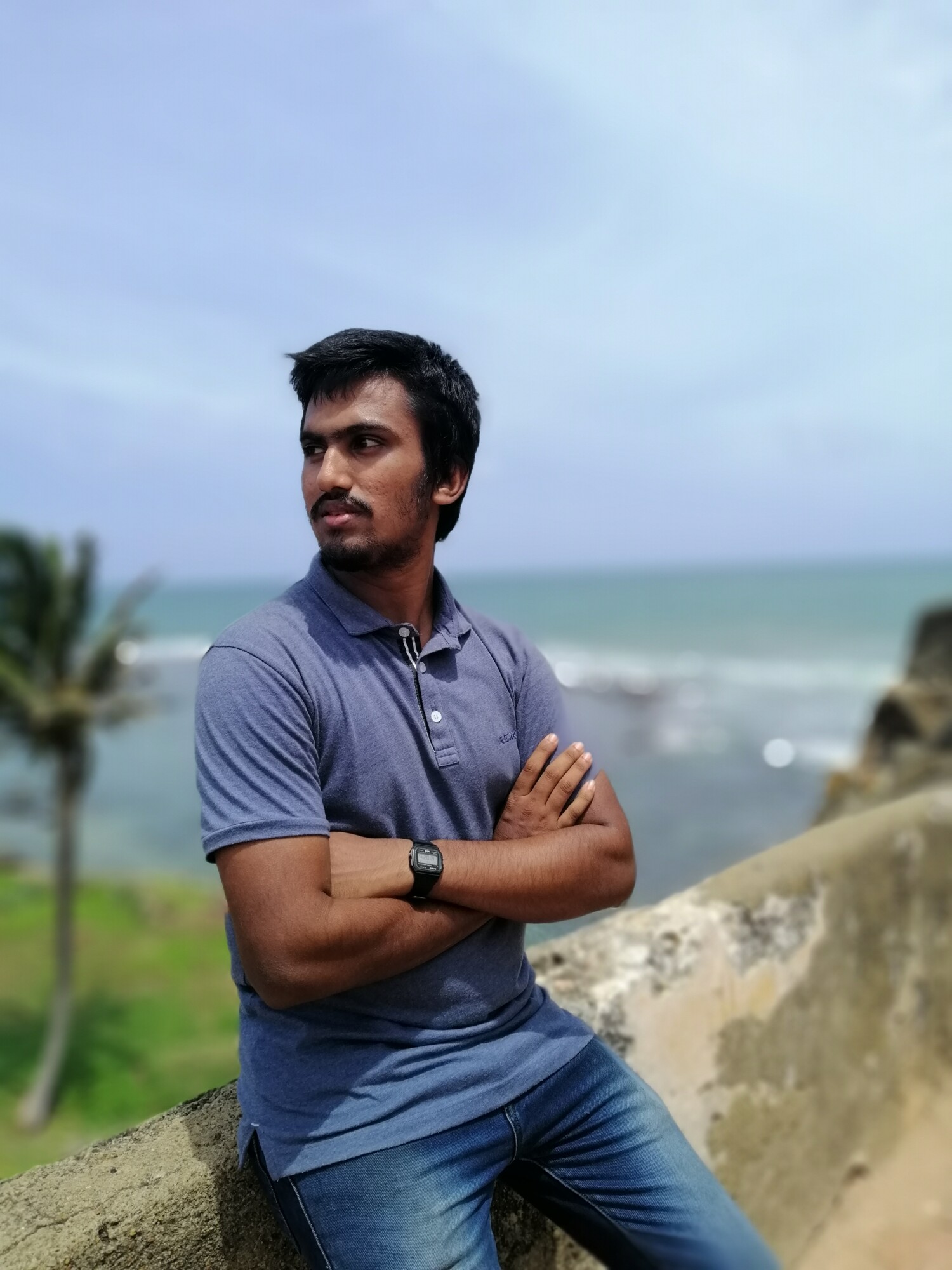 Siniseen poolopaitaan ja farkkuihin pukeutunut mies seisoo kädet ristissä kiviseinää vasten. Taustalla näkyy meri, palmu ja sininen taivas pilvineen.