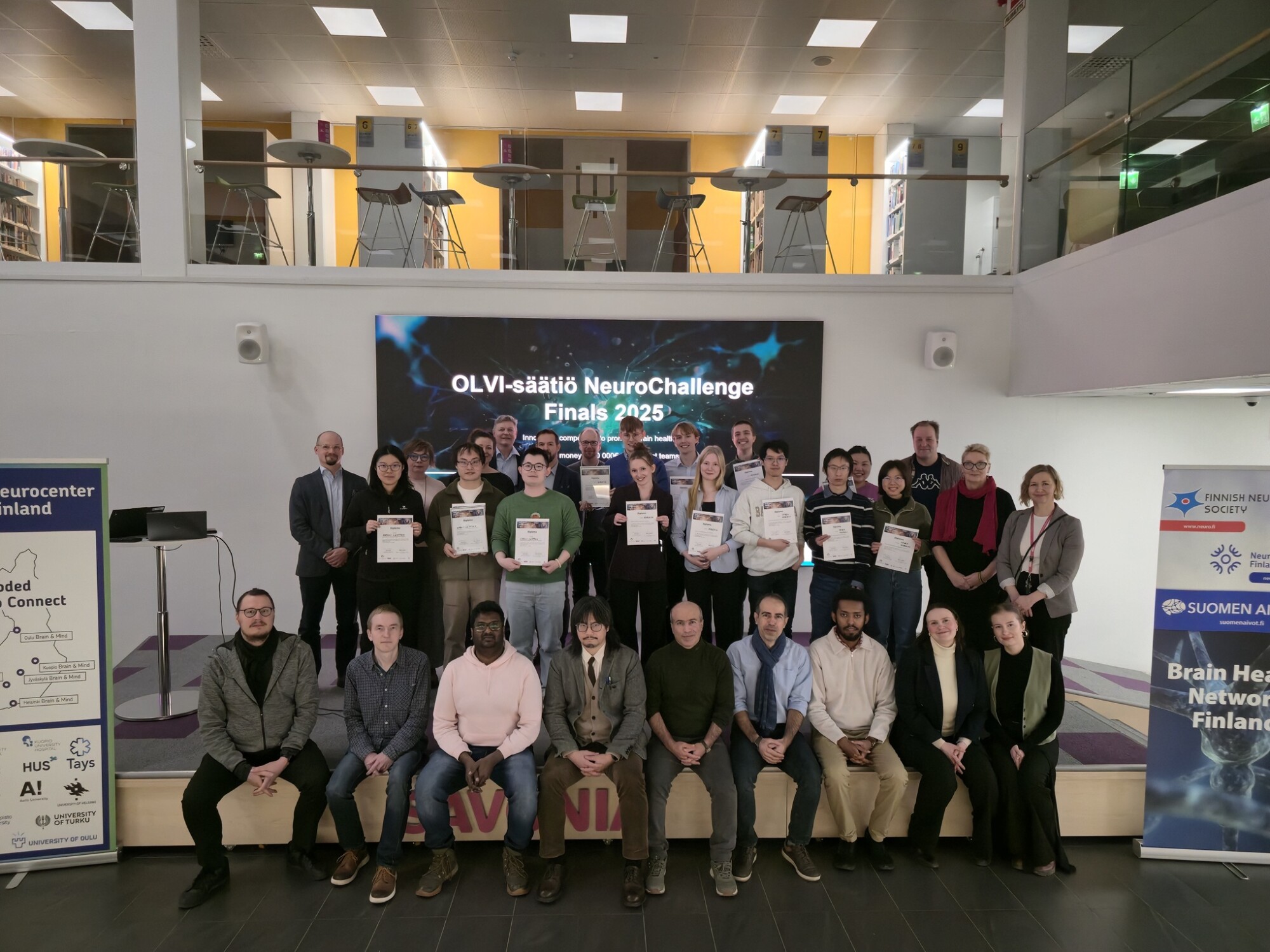 Joukko ihmisiä poseeraa sisätiloissa kolmessa rivissä OLVI-säätiön NeuroChallenge 2023 -finaalissa, ja jotkut pitävät todistuksia kädessään suuren näytön ja tapahtumabanderollien edessä.