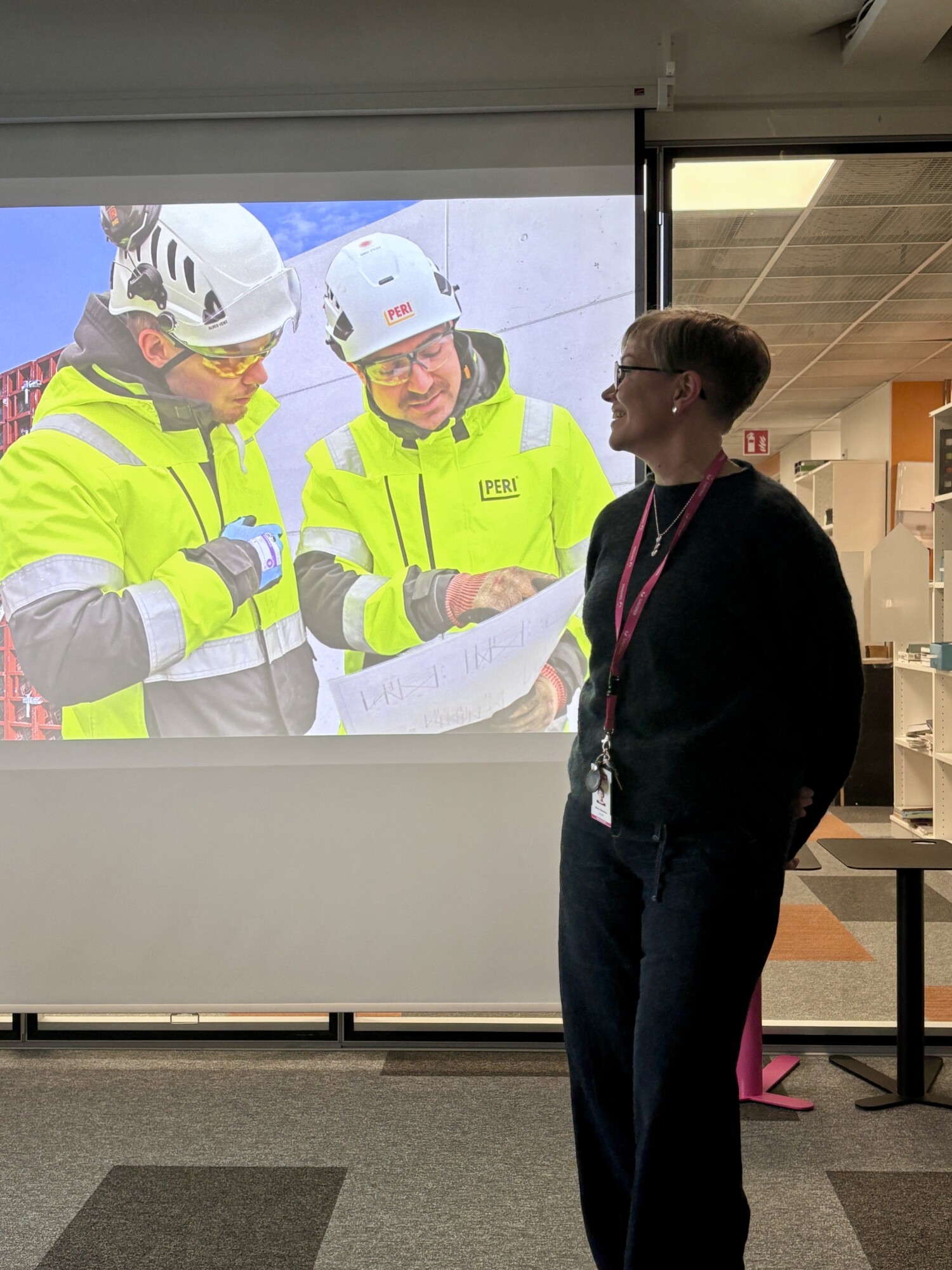 Nainen seisoo ja hymyilee projisoidun kuvan edessä, jossa kaksi turvavarusteisiin pukeutunutta rakennustyöntekijää katselee piirustuksia. Taustalla on toimistokalusteita ja hyllyjä.