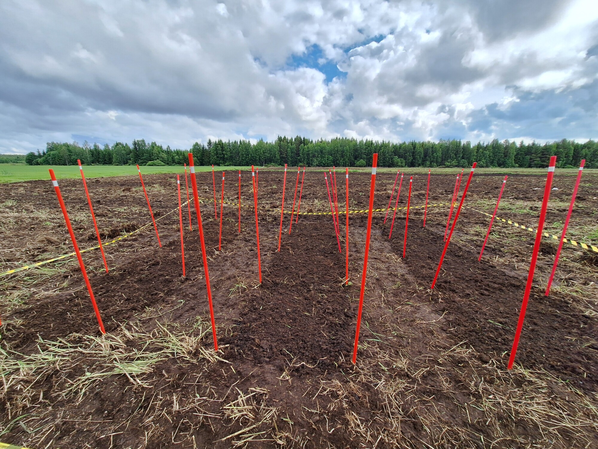 Pelto, jonka maaperään on merkitty punaisilla seipäillä ruudukkoja, joita ympäröi keltainen varoitusnauha, pilvisen taivaan alla, taustalla puiden raja.
