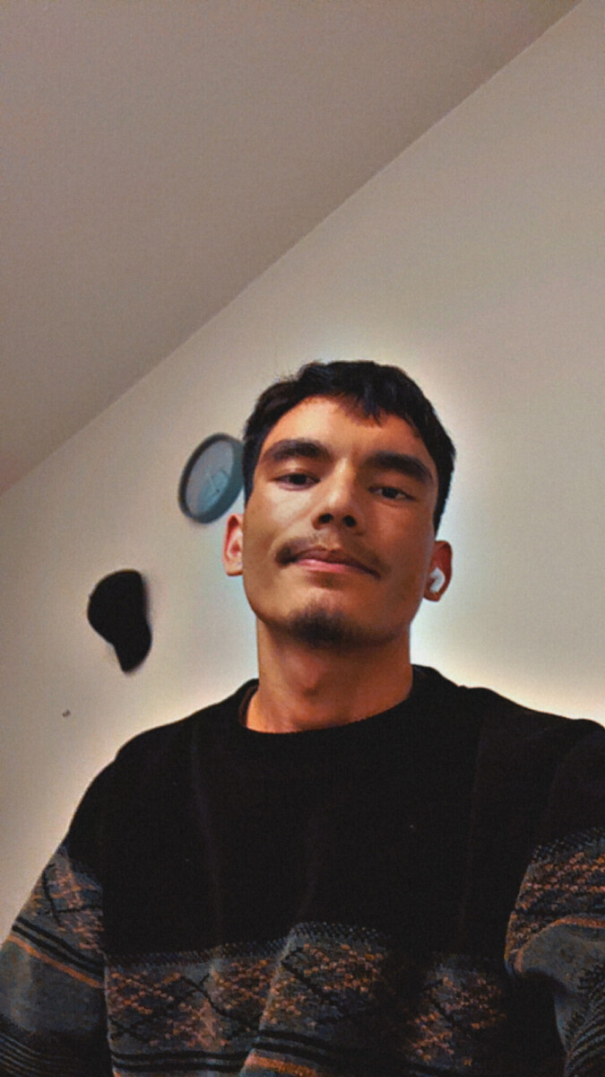 Nuori mies, jolla on lyhyet tummat hiukset ja viikset, hymyilee hieman kameralle. Hänellä on yllään tumma kuviollinen villapaita ja hän istuu sisätiloissa valkoista seinää vasten, ja hänen takanaan roikkuu kello ja musta hattu.